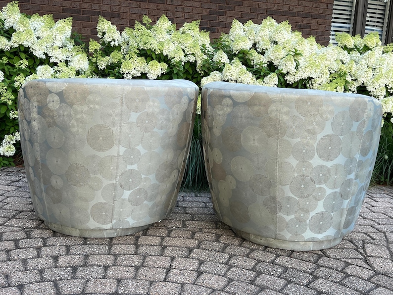 Op de afbeelding: Twee tonvormige fauteuils met een lichtgrijze stof met cirkelpatroon. De stoelen staan op een stenen terras met op de achtergrond witte hortensiastruiken en een bakstenen muur.