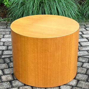 May include: A cylindrical wooden side table with a light brown finish. The table is round and has a smooth surface. The table is approximately 18 inches in diameter and 18 inches tall. The table is made of wood and has a natural wood grain.