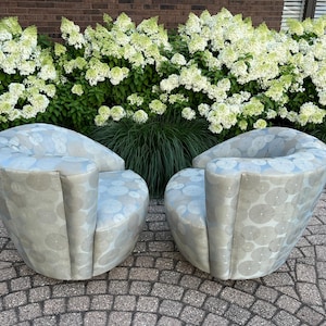 Op de afbeelding: Twee moderne, tonvormige fauteuils met een lichtblauw en grijs cirkelpatroon. De stoelen zijn gestoffeerd en hebben gebogen rugleuningen en armleuningen. Ze staan op een stenen terras voor een haag van witte bloemen.