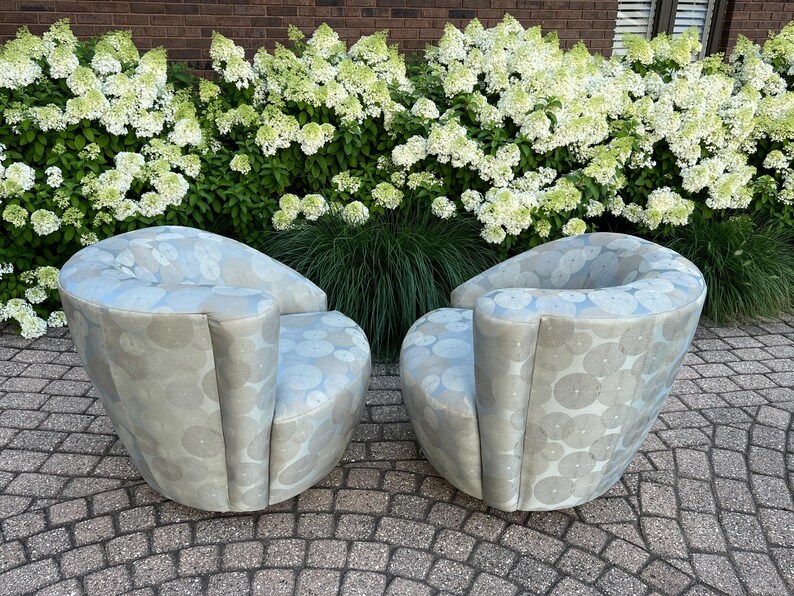 Op de afbeelding: Twee moderne, tonvormige fauteuils met een lichtblauw en grijs cirkelpatroon. De stoelen zijn gestoffeerd en hebben gebogen rugleuningen en armleuningen. Ze staan op een stenen terras voor een haag van witte bloemen.