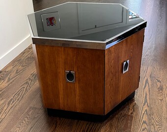 May include: An octagonal wooden coffee table with a dark glass top and metal trim. The table has a rich, brown wood grain finish and two metal ring handles. The base is black.