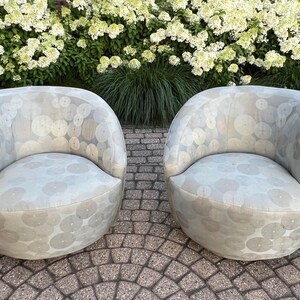 Op de afbeelding: Twee lichtgrijze, afgeronde fauteuils met een cirkelvormig patroon. De stoelen zijn gestoffeerd en hebben een laag profiel. Ze staan op een stenen terras met groene bladeren en witte bloemen op de achtergrond.