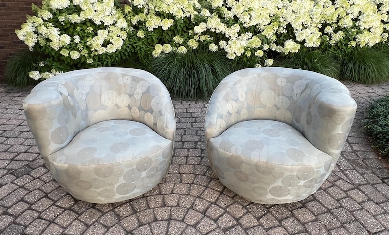 Op de afbeelding: Twee lichtgrijze, afgeronde fauteuils met een cirkelvormig patroon. De stoelen zijn gestoffeerd en hebben een laag profiel. Ze staan op een stenen terras met groene bladeren en witte bloemen op de achtergrond.