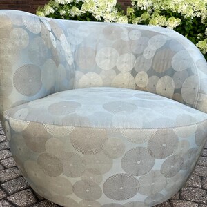 Op de afbeelding: Een lichtblauwe, afgeronde fauteuil met een cirkelvormig patroon in zilver en grijs. De stoel heeft een gebogen rugleuning en armleuningen en is bekleed met een zachte stof. De stoel staat op een stenen terras.