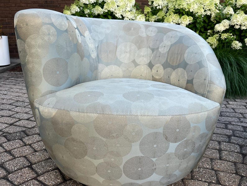 Op de afbeelding: Een lichtblauwe, afgeronde fauteuil met een cirkelvormig patroon in zilver en grijs. De stoel heeft een gebogen rugleuning en armleuningen en is bekleed met een zachte stof. De stoel staat op een stenen terras.