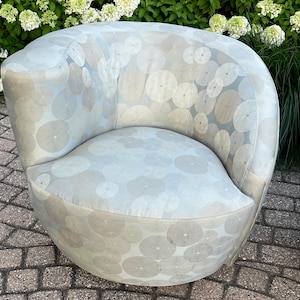 Op de afbeelding: Een lichtblauwe, afgeronde fauteuil met een cirkelvormig patroon in grijstinten en wit. De stoel heeft een gebogen rugleuning en een afgeronde zitting. De stoel is bekleed met een zachte stof en staat buiten.