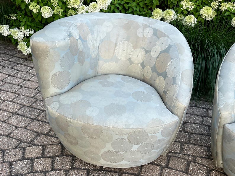 Op de afbeelding: Een lichtblauwe, afgeronde fauteuil met een cirkelvormig patroon in grijstinten en wit. De stoel heeft een gebogen rugleuning en een afgeronde zitting. De stoel is bekleed met een zachte stof en staat buiten.