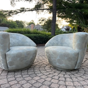 Op de afbeelding: Twee lichtgrijze, afgeronde fauteuils met een cirkelvormig patroon. De stoelen staan op een stenen terras, met groene bladeren en huizen op de achtergrond. De stoelen hebben een modern, gebogen ontwerp.