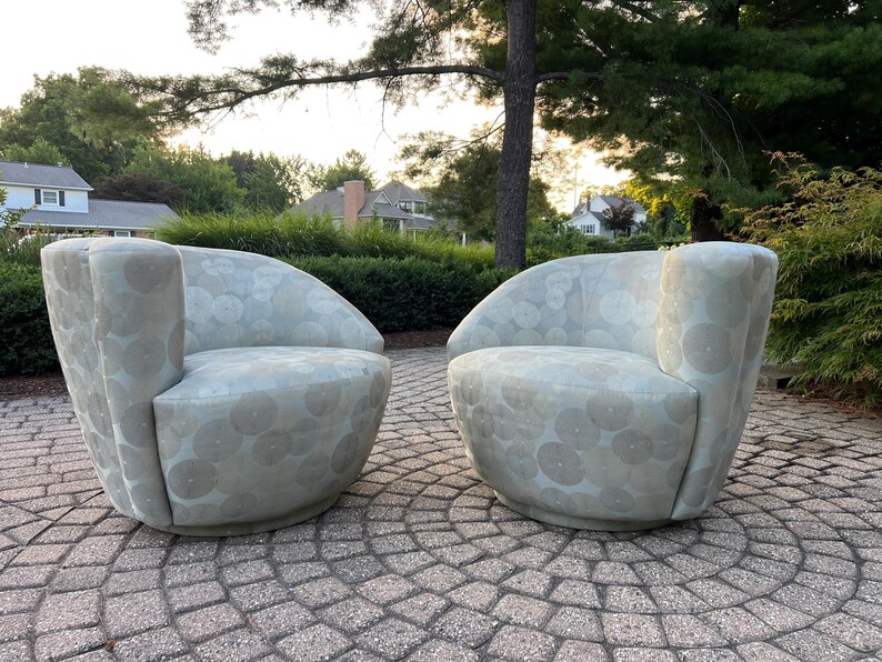 Op de afbeelding: Twee lichtgrijze, afgeronde fauteuils met een cirkelvormig patroon. De stoelen staan op een stenen terras, met groene bladeren en huizen op de achtergrond. De stoelen hebben een modern, gebogen ontwerp.