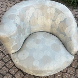 Op de afbeelding: Een lichtblauwe, gebogen fauteuil met een cirkelvormig patroon in grijstinten en beige. De stoel heeft een afgeronde rugleuning en een brede, gebogen zitting. De bekleding is een zachte stof en de stoel lijkt buiten te staan.