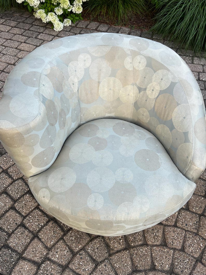 Op de afbeelding: Een lichtblauwe, gebogen fauteuil met een cirkelvormig patroon in grijstinten en beige. De stoel heeft een afgeronde rugleuning en een brede, gebogen zitting. De bekleding is een zachte stof en de stoel lijkt buiten te staan.