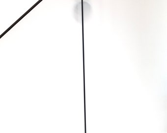 May include: A tall, black floor lamp with a minimalist design. The lamp features a circular base, a slender pole, and a bowl-shaped shade. The lamp is plugged into an electrical outlet.