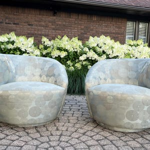 Op de afbeelding: Twee lichtblauwe, tonvormige fauteuils met een cirkelpatroon. De stoelen staan op een stenen terras, met wit en groen bloeiende struiken en een bakstenen muur op de achtergrond.