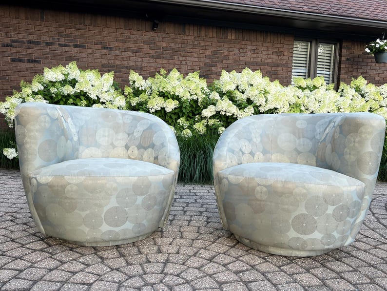 Op de afbeelding: Twee lichtblauwe, tonvormige fauteuils met een cirkelpatroon. De stoelen staan op een stenen terras, met wit en groen bloeiende struiken en een bakstenen muur op de achtergrond.