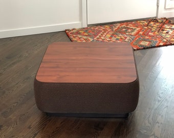 May include: A square ottoman with a smooth, wood-grain patterned top surface and dark brown, textured fabric sides. The piece features rounded corners and sits on a low, dark base, providing a flat surface for use as a footrest or table.