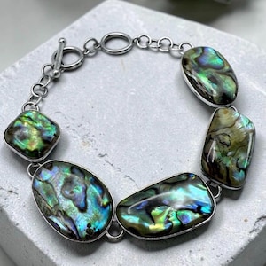 May include: A bracelet featuring five polished, iridescent abalone shell cabochons in varied oval and rectangular shapes, set in silver-toned metal frames and connected by a silver-toned chain with a toggle clasp closure.