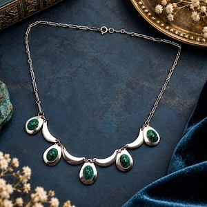 May include: A silver-toned necklace featuring a delicate chain and five teardrop-shaped pendants, each set with a smooth, oval-shaped green stone with dark speckles, interspersed with curved, crescent-like silver metal segments.