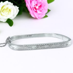 May include: A silver bracelet with a rectangular shape and a floral pattern. The bracelet has a safety chain and is marked with "925". Pink and white roses are in the background.