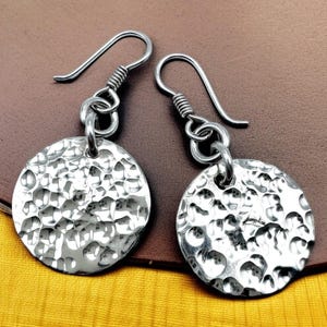 May include: A pair of silver-colored hammered disc earrings. The earrings have a textured surface and are suspended from curved ear wires. The discs are round and have a hammered, dimpled appearance. The earrings are displayed against a brown background.
