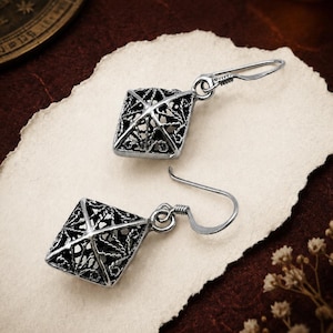 May include: A pair of silver-toned drop earrings featuring a square, pyramid-shaped cage design with intricate, openwork filigree detailing. Each earring is suspended from a simple curved hook wire, showcasing a dark, oxidized finish in the recessed areas.