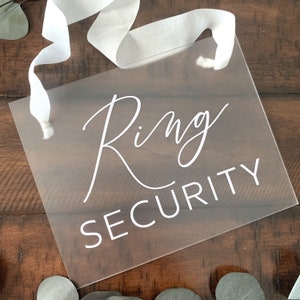 May include: A clear acrylic sign with white text that reads "Ring Security". The sign is hanging from a white ribbon.