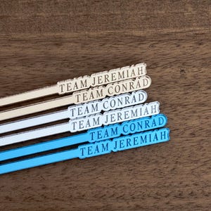 Op de afbeelding: Vier cocktailroerstaafjes met de tekst "TEAM JEREMIAH" en "TEAM CONRAD" in een schreefletter. Twee roerstaafjes zijn gebroken wit en twee lichtblauw. De roerstaafjes liggen op een donker houten oppervlak.