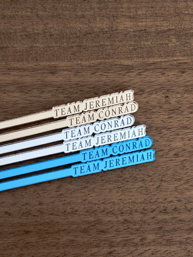 Op de afbeelding: Vier cocktailroerstaafjes met de tekst "TEAM JEREMIAH" en "TEAM CONRAD" in een schreefletter. Twee roerstaafjes zijn gebroken wit en twee lichtblauw. De roerstaafjes liggen op een donker houten oppervlak.