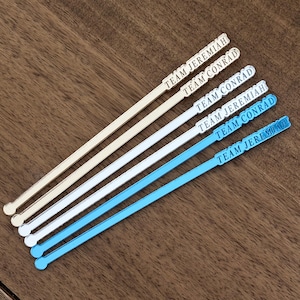 Op de afbeelding: Vijf cocktailroerstaafjes in cr&egrave;me, wit en blauw. Elk roerstaafje heeft een rechthoekige bovenkant met de tekst "TEAM JEREMIAH" of "TEAM CONRAD". De roerstaafjes zijn diagonaal op een houten oppervlak geplaatst.