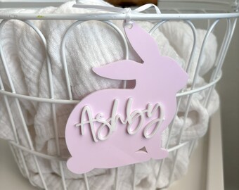 May include: Pink bunny-shaped tag with the name "Ashby" written in white lettering. The tag is hanging from a white wire basket.