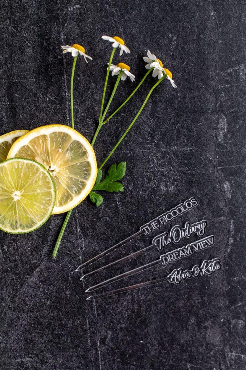 May include: A dark, textured surface with lemon and lime slices, daisy-like flowers, and four clear cocktail stirrers. The stirrers have text that reads "THE PICCOLOS", "The Ordinary", "DREAM VIEW", and "Adam & Katie".