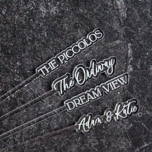 May include: Clear acrylic drink stirrers with white lettering. The stirrers have different text: "THE PICCOLOS", "The Ordinary", "DREAM VIEW", and "Adam & Katie". The background is a dark, textured surface.