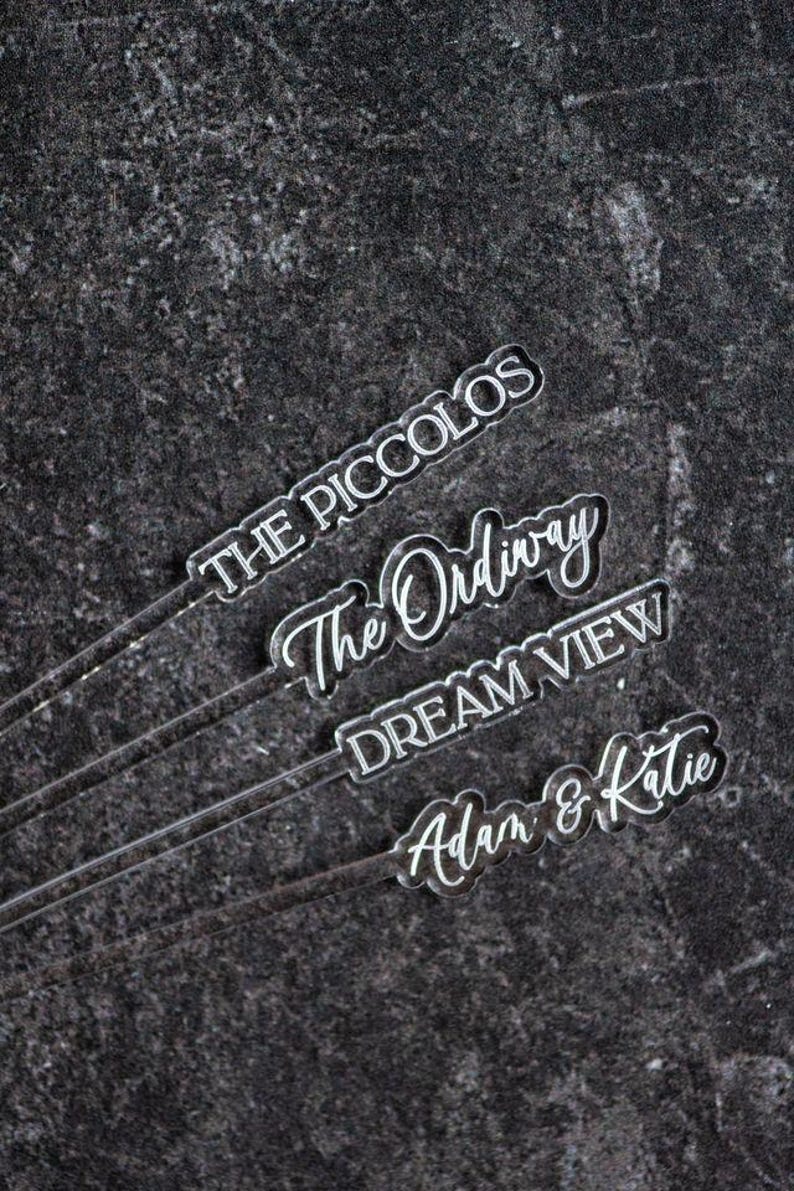 May include: Clear acrylic drink stirrers with white lettering. The stirrers have different text: "THE PICCOLOS", "The Ordinary", "DREAM VIEW", and "Adam & Katie". The background is a dark, textured surface.