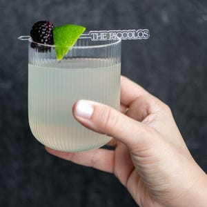 May include: A clear, ribbed glass filled with a light-colored beverage, garnished with a blackberry and lime wedge. A small, clear sign that reads "THE PICCOLOS" is placed on top. The glass is held by a hand against a dark background.