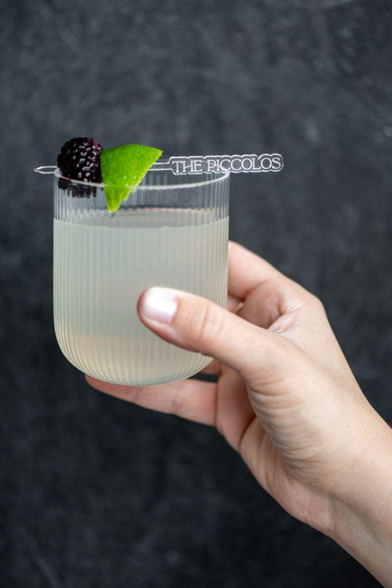 May include: A clear, ribbed glass filled with a light-colored beverage, garnished with a blackberry and lime wedge. A small, clear sign that reads "THE PICCOLOS" is placed on top. The glass is held by a hand against a dark background.