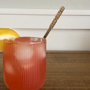 Op de afbeelding: Een verfrissende roze cocktail in een geribbeld glas met ijsblokjes en een citroenpartje. Een goudkleurige barlepel met de tekst "TEAM CONRAD" zit in de drank. De drank staat op een houten oppervlak.