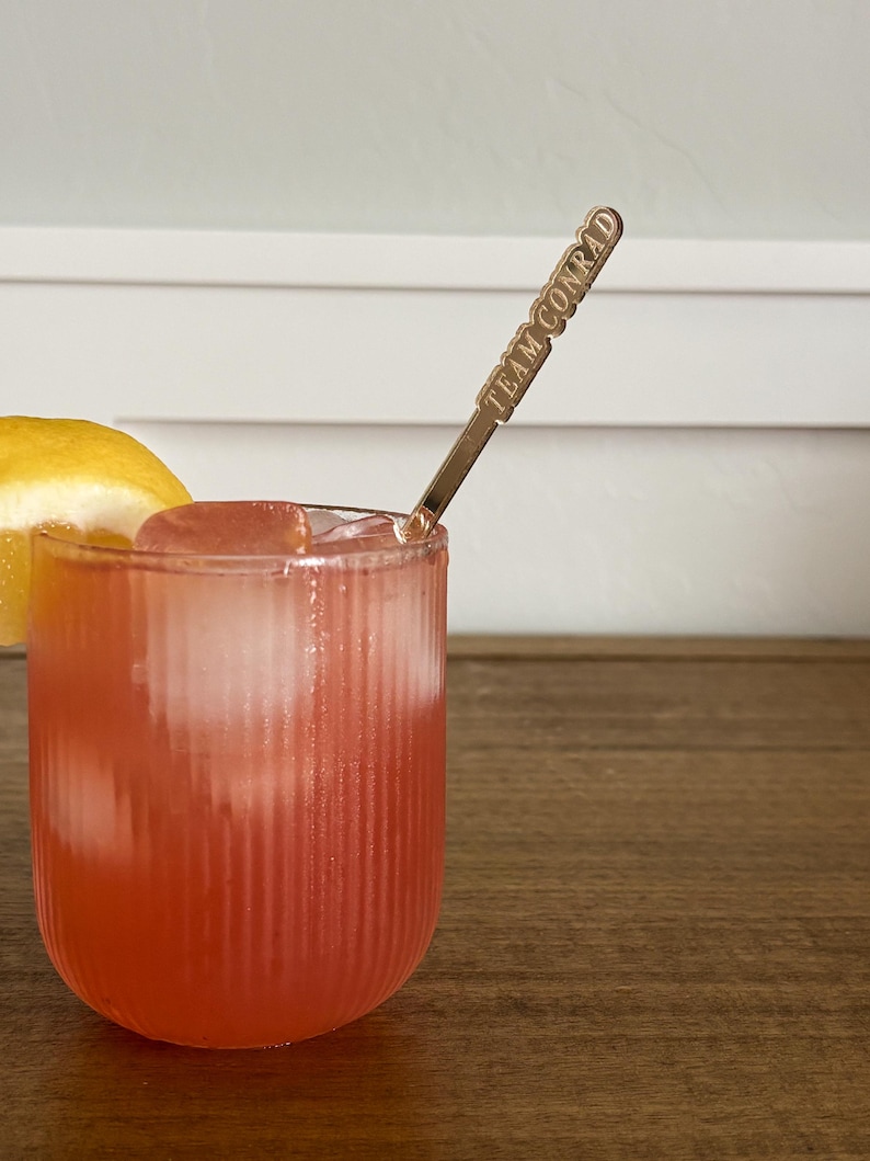 Op de afbeelding: Een verfrissende roze cocktail in een geribbeld glas met ijsblokjes en een citroenpartje. Een goudkleurige barlepel met de tekst "TEAM CONRAD" zit in de drank. De drank staat op een houten oppervlak.