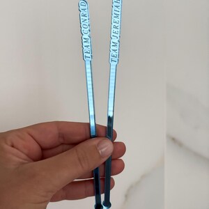 Op de afbeelding: Twee blauwe cocktailroerstaafjes met witte tekst. E&eacute;n leest "TEAM CONRAD", de andere "TEAM JEREMIAH". De roerstaafjes zijn dun, rechthoekig en hebben een kleine cirkelvormige basis. De achtergrond is licht en neutraal.