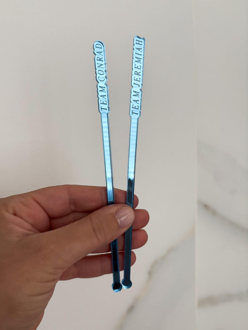 Op de afbeelding: Twee blauwe cocktailroerstaafjes met witte tekst. E&eacute;n leest "TEAM CONRAD", de andere "TEAM JEREMIAH". De roerstaafjes zijn dun, rechthoekig en hebben een kleine cirkelvormige basis. De achtergrond is licht en neutraal.