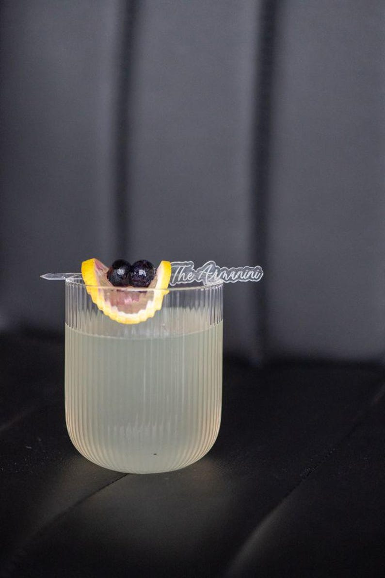 May include: A refreshing cocktail in a ribbed glass, garnished with a lemon slice and dark berries. A clear plastic stirrer with the text "The Arannini" rests on the rim. The drink is set against a dark background.