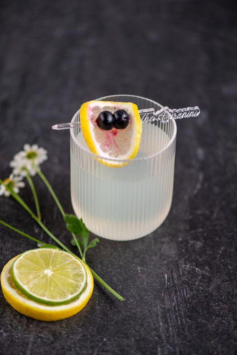 May include: A refreshing cocktail in a ribbed glass, garnished with a lemon slice and two dark berries. A lime and lemon slice are at the bottom left. The cocktail is on a dark surface.