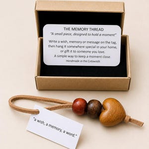 Op de afbeelding: Een houten hartvormig aandenken met een leren lus en houten kralen, gepresenteerd in een kartonnen doos. Het label luidt "a wish, a memory, a word." De doos bevat een kaart met tekst.