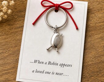 May include: A silver-colored bird keychain with a red bow, attached to a white card with the text "...When a Robin appears a loved one is near...." The keychain is on a wooden surface.