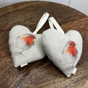 May include: Two heart-shaped fabric ornaments, each with a robin illustration. The robins have red breasts and are set against a beige background. The ornaments have a textured finish and cream ribbon loops for hanging. The fabric has a speckled appearance.