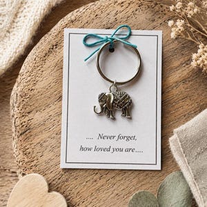 Peut inclure: Porte-clés éléphant argenté avec un motif décoratif, attaché à une carte blanche avec le texte "... Never forget, how loved you are...". La carte est attachée avec une ficelle turquoise. Le porte-clés est sur une surface en bois.