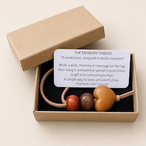 Op de afbeelding: Een geheugendraadset in een kartonnen doos. De set bevat een leren koord met drie houten kralen en een hartvormige bedel. Een kaart met de tekst "THE MEMORY THREAD" en instructies. Handgemaakt in de Cotswolds.