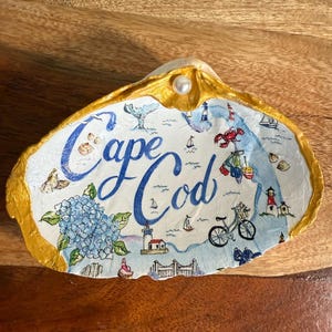 May include: A decorative oyster shell featuring the text Cape Cod in blue script. The shell is painted with illustrations of blue hydrangeas, lighthouses, a bicycle, a lobster, and sailboats. The edges are painted gold, and a small white pearl is attached.
