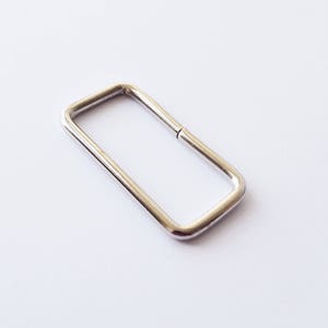 May include: A silver metal rectangular ring or loop, often used for attaching straps or other hardware to bags, belts, or other items.