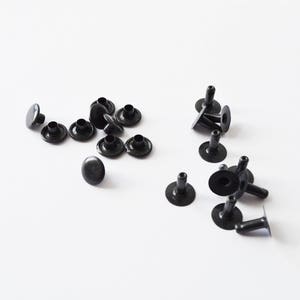 Puede incluir: Primer plano de remaches de metal negro, algunos con poste y otros sin. Los remaches están esparcidos sobre una superficie blanca.