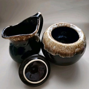 May include: A vintage ceramic creamer and sugar bowl set. The creamer has a handle and spout, while the sugar bowl has a lid. Both pieces are dark brown with a lighter brown rim. The lid is off the sugar bowl, revealing the interior.