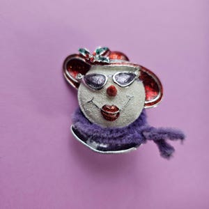 May include: A white snowman ornament with a red hat, purple scarf, and sunglasses. The snowman has a red glitter mouth and a white glitter face.